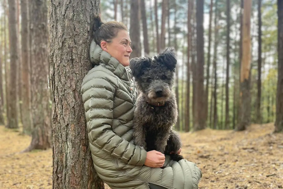 Kvinna lutar sig med ryggen mot ett träd och håller en hund i famnen